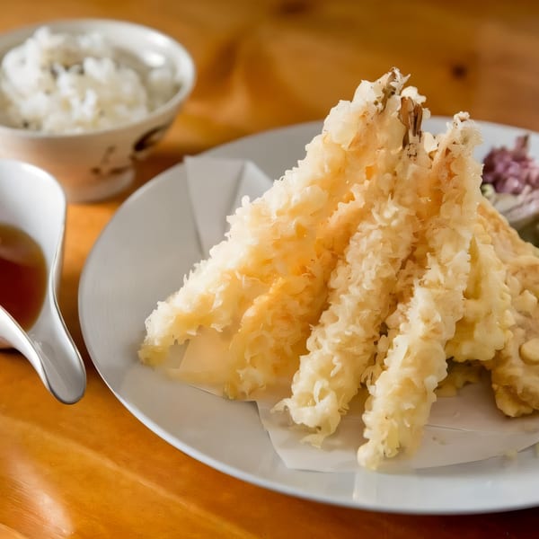 Crispy golden tempura shrimp and vegetables on a white plate with rice and dipping sauce.