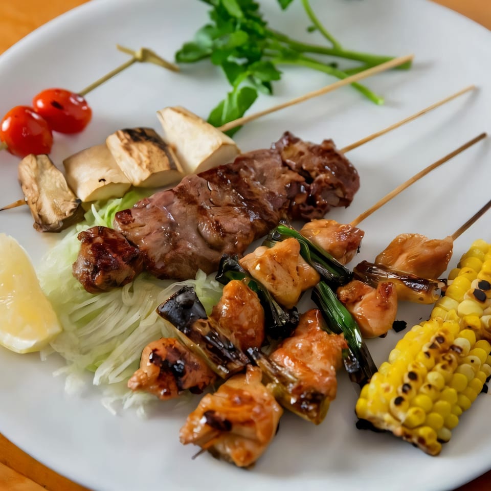 Assortment of Japanese yakitori skewers with chicken, beef, mushrooms, tomatoes, and corn on a white plate.