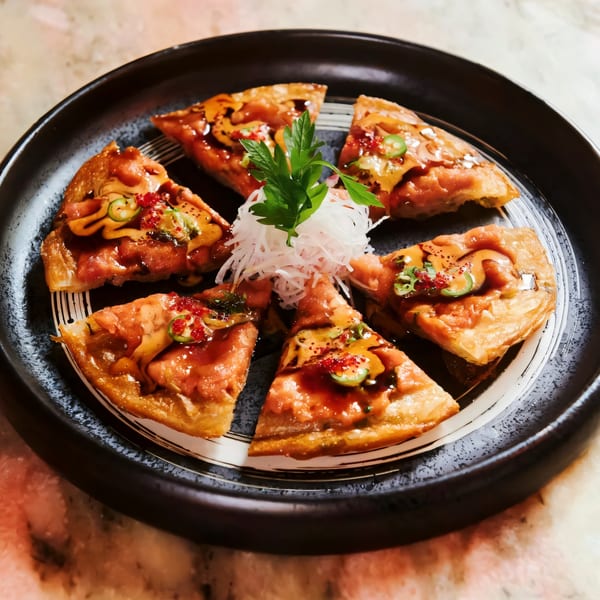 Sapps Nyc's Japanese tuna pizza, cut into six slices, garnished with herbs and sauces on a black plate.
