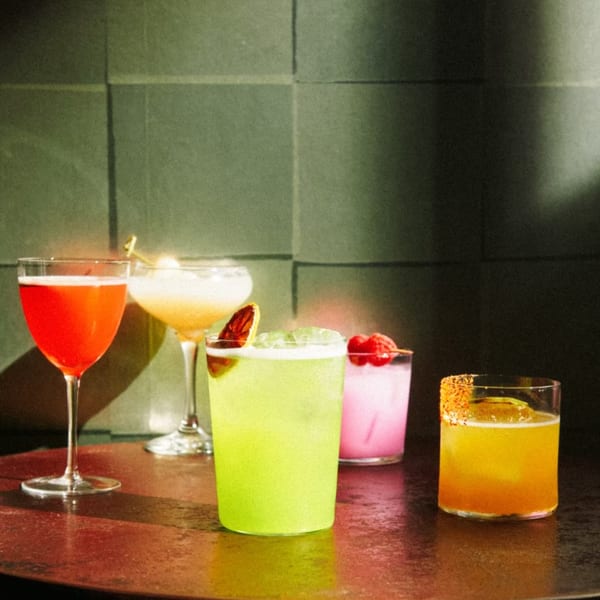Four colorful Sapps NYC cocktails, including red, yellow, green, and pink drinks, on a dark table.
