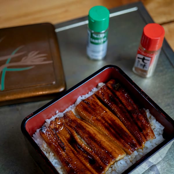 Unagi donburi, grilled eel over rice, in a black and red lacquer box with spice shakers.
