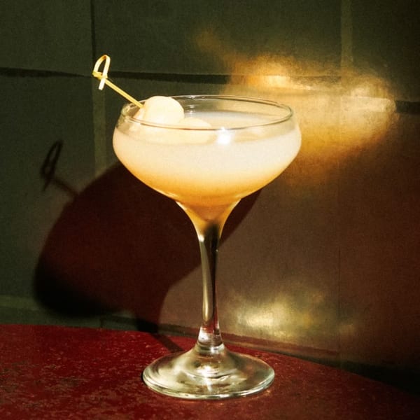 A light-colored cocktail in a coupe glass, garnished with two small white spheres on a pick.