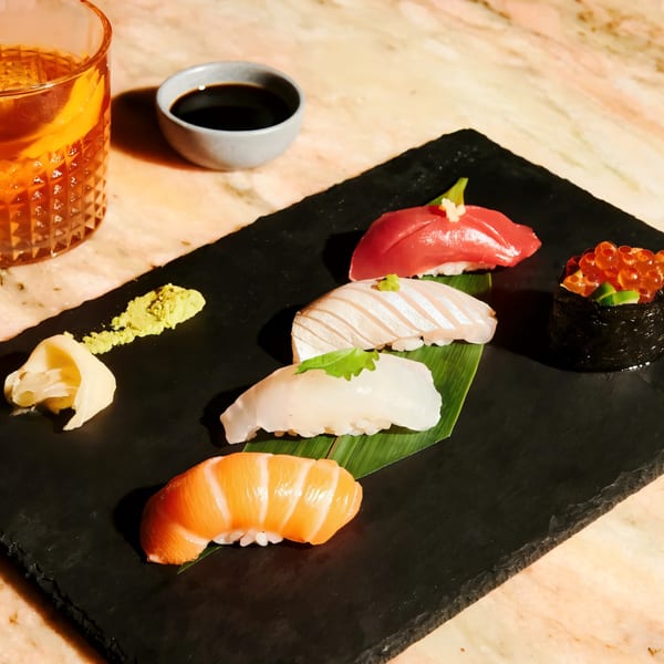 Assortment of Sapps Nyc sushi and sashimi on a black slate, with a drink and soy sauce.